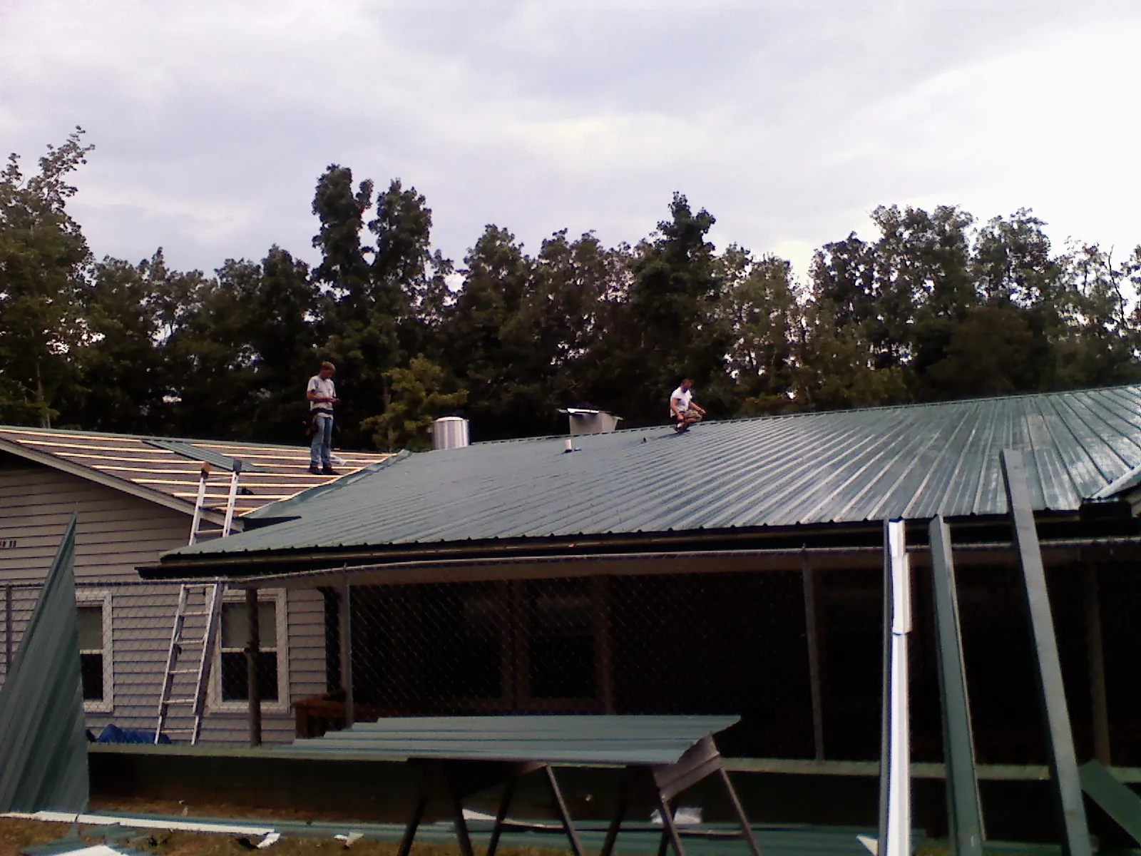 Two roofers installing green metal panels above a covered porch in Dexter